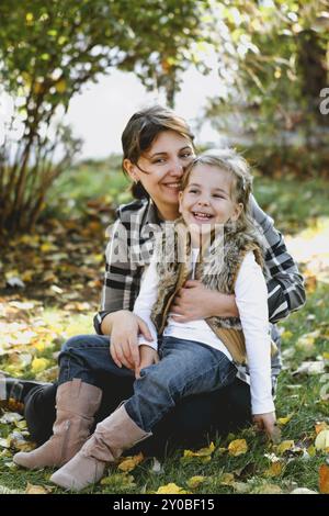 Madre amorevole e ragazza affascinante in abiti caldi abbracciando felicemente seduto su prato verde con foglie cadute alla luce del sole Foto Stock