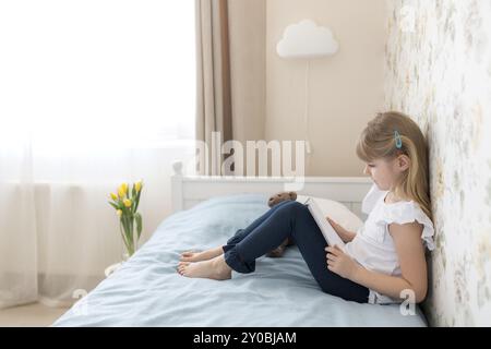 Una bambina si siede sul letto nell'elegante camera da letto e legge un libro blu. Istruzione, concetto di scuola domestica. Fare i compiti. Tulipani gialli nel va Foto Stock