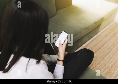 Concetto di comunicazione con Black Hear ragazza seduta su un divano verde e tenendo lo smartphone in mano con schermo bianco vuoto, fascio di sole sulla parte anteriore Foto Stock