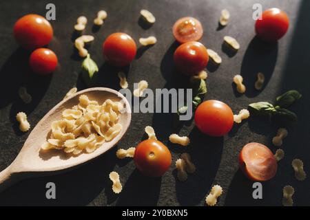 Pomodori ciliegini rossi, basilico verde, vermicelli a farfalla, cucchiaio di legno su sfondo grigio. Foto Stock