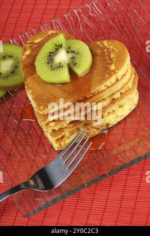 Pila di pancake a forma di cuore con sciroppo e kiwi su un piatto di vetro trasparente su sfondo rosso. Una colazione perfetta per San Valentino Foto Stock