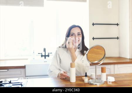 Carino donna sorridente l'applicazione di crema per il viso Indossare tunica bianca al mattino presso la cucina Foto Stock