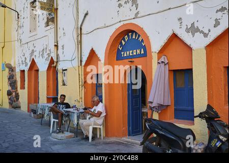 Persone sedute di fronte a una facciata colorata di una taverna e godendosi il tempo libero per strada, Mandraki, Nisiros, Dodecaneso, Isole greche, Grecia Foto Stock