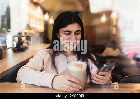 Ragazza sorridente, beve caffè al bar e legga il telefono. Sfondo sfocato. Foto Stock