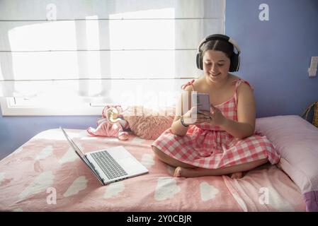 Una ragazza con un abito a scacchi si siede su un letto rosa con le cuffie, tenendo un telefono e un portatile accanto a lei in una camera accogliente e luminosa, tarragona, spagna Foto Stock