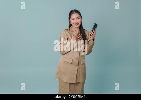 Felice consapevole grato indonesiano lavoratrice del governo mano sul petto sorridente mentre tiene il telefono. Concetto di funzionario pubblico PNS. Foto Stock