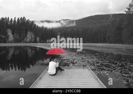 Una foto concettuale di un'immagine in bianco e nero con una donna che regge un ombrello rosso mentre è seduta su un molo vicino a un lago nell'Idaho settentrionale Foto Stock