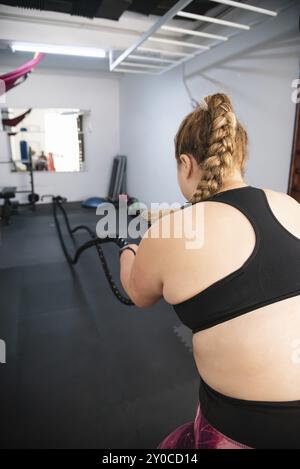 Una giovane donna, piena di dedizione, si impegna in un'intensa sessione di allenamento con corde di battaglia nell'ambiente dinamico di una palestra ben attrezzata Foto Stock