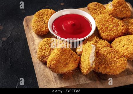 Pepite di pollo con ketchup primo piano su un asse di legno. Uno spuntino croccante in un ristorante, un piatto fast food su sfondo nero Foto Stock