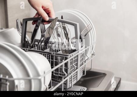 Primo piano della mano femmina che tiene il cestello completo delle posate con coltello pulito, forchetta, frusta. Caricamento, svuotamento o scaricamento dalla lavastoviglie automatica aperta m Foto Stock