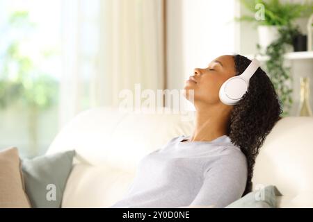 Profilo di una donna nera che indossa le cuffie respirando e ascoltando musica seduta sul divano di casa Foto Stock