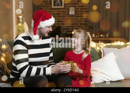 Buon Natale e Felice Anno Nuovo. Sorpresa regalo. Una ragazza sorridente piccola felice apre un regalo di Natale in stupore. Padre dà il suo malessere Foto Stock