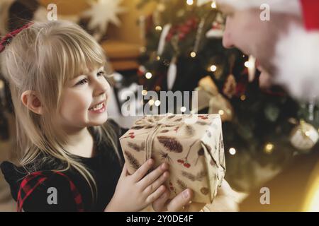 Giovane padre in cappello rosso di Santa che dà il regalo di Natale alla sua figlia sorridente vicino decorato albero di Natale. Ragazza vestita di Natale rosso-nero festivo Foto Stock