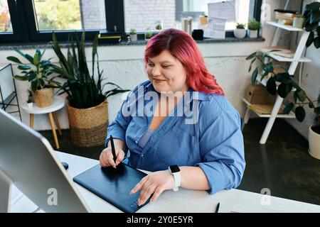 Una donna di grandi dimensioni con i capelli rossi accesi disegna su un tablet digitale mentre lavora alla scrivania. Foto Stock