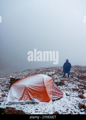 Un escursionista con una tenda in cima alle montagne in una nevicata. Uomo barbuto in impermeabile blu e tenda rossa durante le nevicate Foto Stock