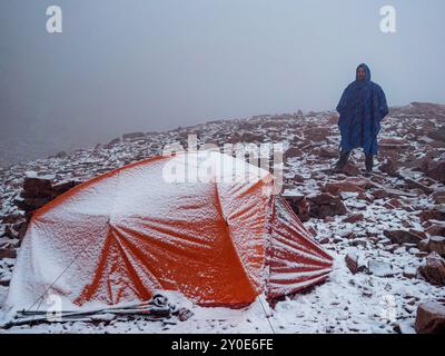 Un escursionista con una tenda in cima alle montagne in una nevicata. Uomo barbuto in impermeabile blu e tenda rossa durante le nevicate Foto Stock