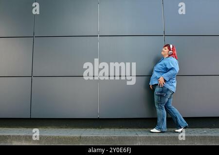 Una donna più grande in jeans blu e una camicia blu passa davanti a un muro grigio. Foto Stock