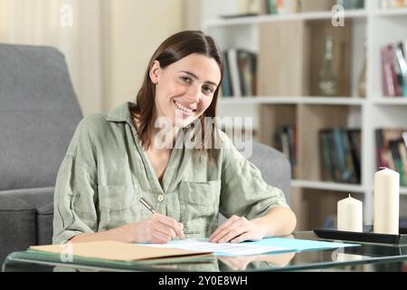 Donna felice che firma contratti e guarda la macchina fotografica seduta a casa Foto Stock