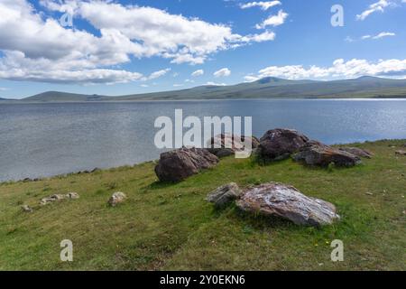 Parte occidentale del lago Paravani, rocce ricoperte di licheni arancioni vicino alla riva. Catena montuosa coperta da nuvole Foto Stock