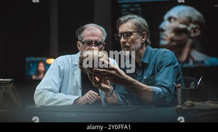 Due archeologi effettuano misurazioni del cranio umano preistorico, conducono ricerche scientifiche, usano un tablet. Gli scienziati esaminano i resti fossili in un laboratorio archeologico avanzato di notte. Foto Stock