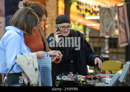 Tbilisi, Georgia - 8 ottobre 2023: Due donne sfogliano gioielli fatti a mano mentre un venditore anziano parla al telefono. Foto Stock