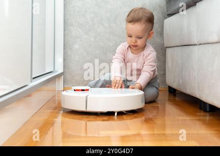 Una bambina curiosa che gioca con l'aspirapolvere robot sul pavimento in parquet giallo Foto Stock