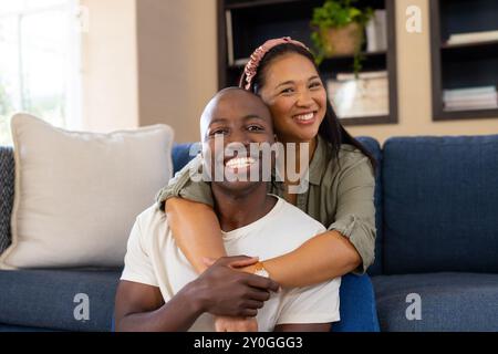 Coppia multirazziale sorridente seduta sul divano, abbracciando e divertendosi a casa Foto Stock