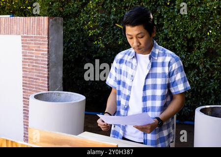 Revisione dei progetti, uomo asiatico in camicia a scacchi che lavora su progetti di costruzione all'aperto Foto Stock