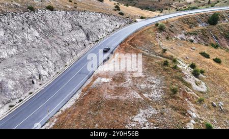 Un'auto solitaria percorre una strada tortuosa attraverso un terreno collinare durante una giornata limpida in un paesaggio montuoso Foto Stock