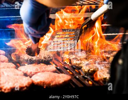 Cottura di hamburger su grill caldo con fiamme - pinze e spatola che muovono polpette grigliate alla fiamma e crude di hamburger alla griglia. Gli hamburger sono tradizionali Foto Stock