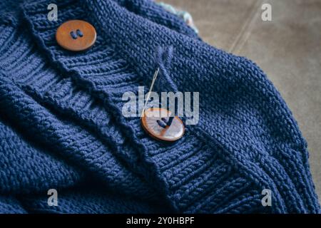 Cardigan in maglia di lana blu con bottone in legno e ago da cucire. Unione del modello di nervatura con i bottoni. Cardigan fatto a mano. Sfondo abiti caldi. Foto Stock