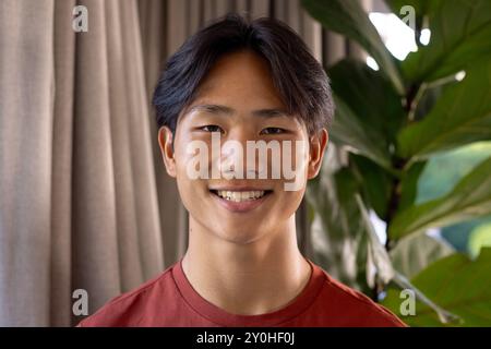 Sorridente giovane asiatico in camicia rossa in posa in interni con pianta verde sullo sfondo Foto Stock