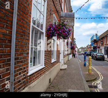 Vista sulla strada di un'affascinante città con cesti di fiori appesi, edifici in mattoni e alcune auto parcheggiate lungo il lato . Winchester Regno Unito Foto Stock