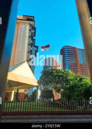 Bandiera indonesiana che sventola su un cielo azzurro con un moderno edificio per uffici sullo sfondo. Foto Stock