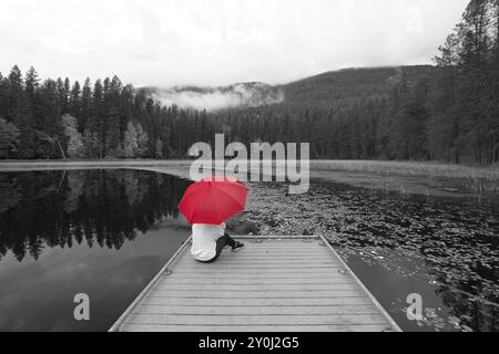 Una foto concettuale di un'immagine in bianco e nero con una donna che regge un ombrello rosso mentre è seduta su un molo vicino a un lago nell'Idaho settentrionale Foto Stock