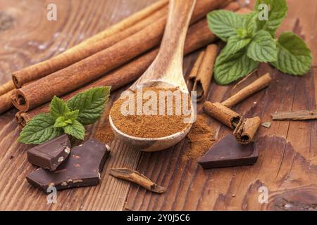 Polvere di cannella su un cucchiaio di legno, foglie di menta, bastoncini di cannella e pezzi di cioccolato fondente Foto Stock