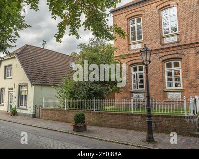 Tranquilla strada rurale con una grande casa in mattoni, un albero e una lanterna sotto un cielo nuvoloso, dornum, frisia orientale, germania Foto Stock