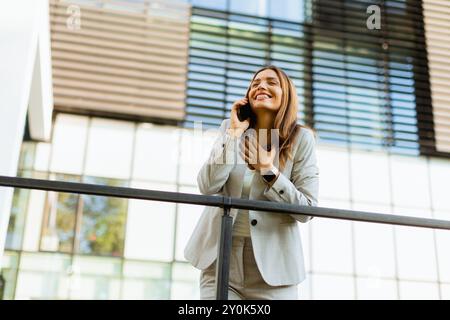 Una donna d'affari gioiosa festeggia un momento all'aperto, partecipando a una allegra conversazione telefonica mentre si appoggia a una ringhiera accanto a un elegante buildi Foto Stock