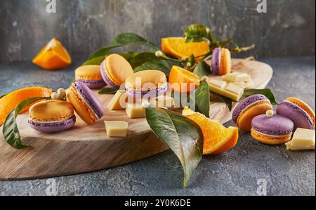Vivaci macaron gialli e viola visualizzati con fette arancioni, foglie verdi e pezzi di cioccolato bianco su una tavola rustica di legno. Foto Stock