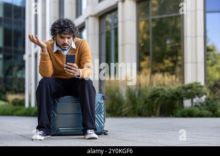 Un giovane che guarda il telefono con confusione seduto sulla valigia fuori dal moderno edificio per uffici. I professionisti aziendali sembrano frustrati durante l'attesa, bilanciando l'utilizzo della tecnologia in ambienti urbani. Foto Stock