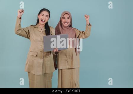 Due gioiose lavoratrici governative che indossano l'uniforme cachi e tengono un computer portatile con un'espressione felice di successo. PNS e concetto di funzionario pubblico. Foto Stock