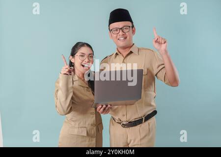 Una coppia di dipendenti governativi entusiasta in uniforme cachi che tiene un laptop mentre punta verso l'alto lo spazio di copia. PNS e concetto di funzionario pubblico. Foto Stock
