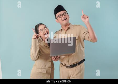 Una coppia di dipendenti governativi entusiasta in uniforme cachi che tiene un laptop mentre punta verso l'alto lo spazio di copia. PNS e concetto di funzionario pubblico. Foto Stock