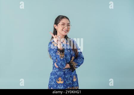 Allegra lavoratrice governativa in uniforme blu batik e occhiali che puntano allo spazio di copia dietro di lei con un sorriso. Concetto di funzionario pubblico PNS. Foto Stock