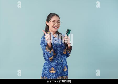 Lavoratrice del governo felice in uniforme blu batik e occhiali che tengono in mano uno smartphone, puntando allo spazio di copia dietro di lei. Dipendenti pubblici PNS co Foto Stock