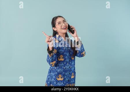 Allegra lavoratrice del governo in uniforme blu batik e occhiali durante una chiamata di lavoro, mentre indica di copiare lo spazio dietro di lei. Concetto di funzionario pubblico PNS. Foto Stock