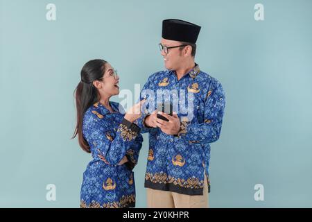 Due gioiosi operai del governo indonesiano che indossano l'uniforme blu batik e occhiali che indicano il cellulare che tiene. Concetto di funzionario pubblico PNS. Foto Stock