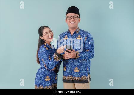 Due gioiosi operai del governo indonesiano che indossano l'uniforme blu batik e occhiali che indicano il cellulare che tiene. Concetto di funzionario pubblico PNS. Foto Stock