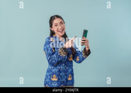Allegra lavoratrice del governo indonesiano in uniforme blu batik e occhiali che indicano il cellulare che ha in mano. Concetto di funzionario pubblico PNS. Foto Stock