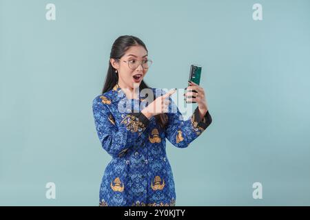 Lavoratrice governativa indonesiana in uniforme blu batik e occhiali che indicano il cellulare che tiene con un look sorpreso. PNS cep. Funzionario pubblico Foto Stock